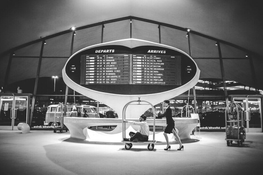 A flight attendant pushes Roman Freud on a luggage cart under an airport departures board