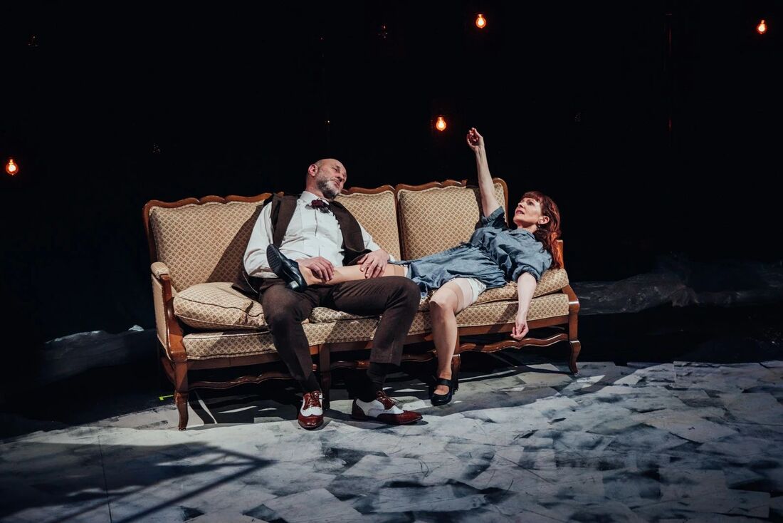 Two actors on a sofa under hanging bulbs