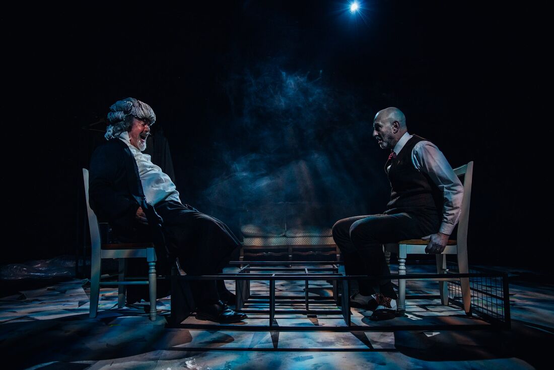 Two men face each other on chairs in blue stage light